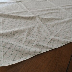 May include: A white fabric tablecloth with a green diamond pattern. The tablecloth is folded and lying on a wooden surface.