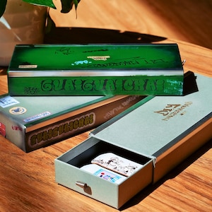 Puede incluir: Caja rectangular verde y plateada con la palabra "GUAGUAICHAI" en letras grandes. La caja está encima de otra caja con las palabras "GUAGUAICHAS" y "DIY FINGERBOARDS". Una tercera caja está abierta, revelando pequeños monopatines.