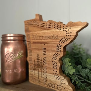 May include: A wooden cribbage board in the shape of the state of Minnesota, with the word "Minnesota" engraved on it. The board features a tree design and numerous holes for pegs. A copper-colored Ball jar is visible in the background.