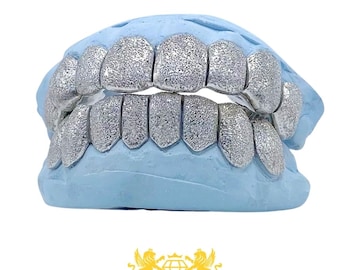 May include: A set of silver-toned, textured grillz fitted onto a light blue dental mould. The grillz cover both the upper and lower teeth, featuring a granular, sparkling surface finish that catches the light across each individual tooth section.