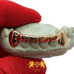 Custom 6-tooth rose gold grillz set with solid fangs and connecting horizontal bar held in red gloved hand