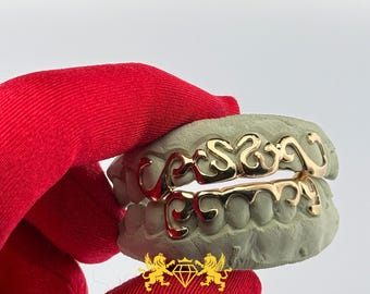 Op de afbeelding: Gouden, op maat gemaakte grillz met de naam "Cason" in een cursief lettertype. De grillz worden getoond op een grijze tandafdruk, vastgehouden door een rode handschoen. Onderaan staat een logo met twee leeuwen en een diamant.