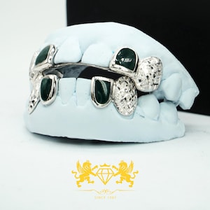 May include: Silver-colored grillz with dark green inlays and textured detailing, displayed on a light blue dental mold. The grillz are custom-fitted and feature a polished finish. The logo "SINCE 1987" is visible.