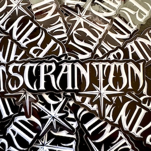 May include: A collection of black and white stickers. Each sticker features the word "SCRANTON" in a stylized font, with a starburst design. The stickers have a glossy finish and are scattered across the image.