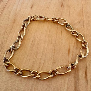 May include: A gold-toned metal chain bracelet featuring elongated, oval-shaped interlocking links. The bracelet is arranged in a circular shape and includes a small, circular spring-ring clasp mechanism at the top of the chain.