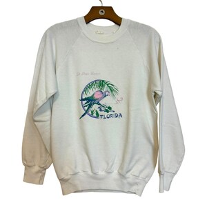 May include: White sweatshirt with a crew neck and long sleeves. The front features a colorful graphic of a parrot perched on a branch, with the words "St. Pete Beach" and "FLORIDA" in a circular design.