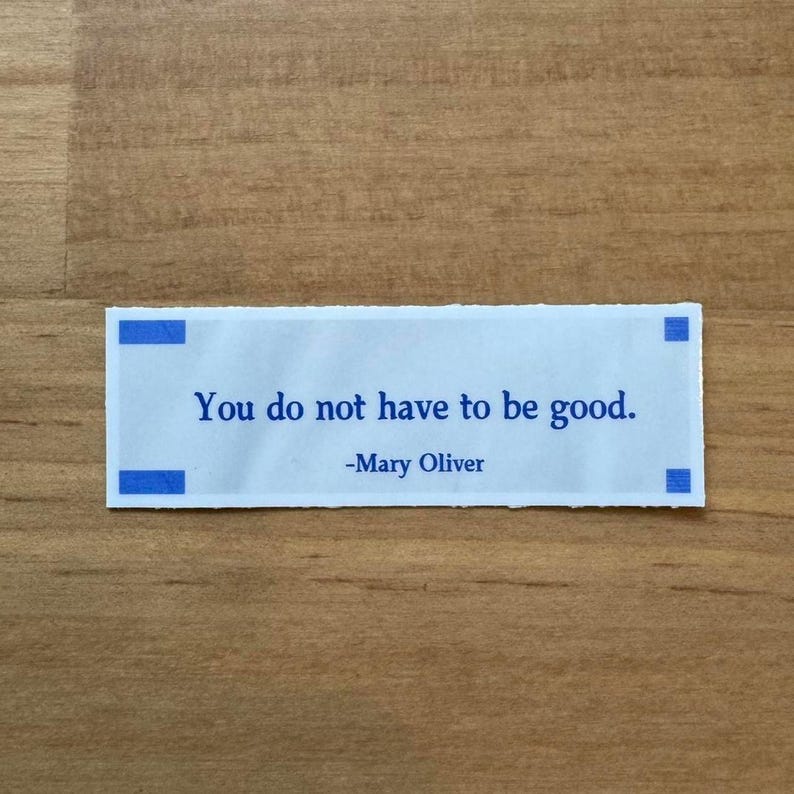 May include: A white fortune cookie message with blue detailing, featuring the quote "You do not have to be good." by Mary Oliver. The message is set against a wooden surface.