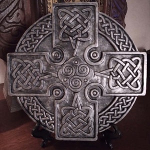 May include: A silver-colored Celtic cross with intricate knotwork designs. The cross is round with a square cross shape in the center. The design includes a central spiral and knot patterns.