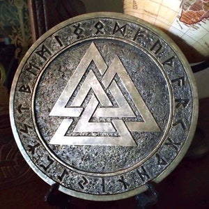 Könnte beinhalten: Eine runde Wanddekoration aus Metall mit einem dreidimensionalen, silberfarbenen, ineinandergreifenden Dreiecksmuster. Das Muster ist von einem Kreis aus schwarzen Runen umgeben.