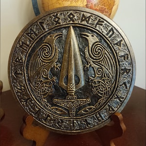 May include: A round, bronze-coloured metal plaque with a raised design of two ravens flanking a spear. The plaque is surrounded by a circle of runes and has a wooden stand.