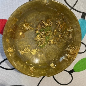 Pode incluir: Um cinzeiro redondo de cor dourada com uma superfície texturizada e flocos dourados embutidos na resina. O cinzeiro tem uma ligeira borda ao redor da borda.