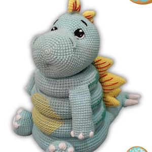 May include: A crocheted dinosaur toy in shades of blue, yellow, and white. The dinosaur has a friendly expression and the word "Fatosaurus" is written above it. Donuts with blue frosting are in the background.
