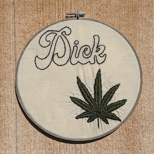 May include: Embroidered artwork in a round hoop. The word "Sick" is stitched in black cursive above a detailed green cannabis leaf. The fabric is a light beige, and the hoop is silver.