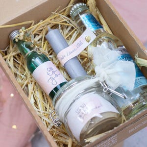 Puede incluir: Una caja de regalo llena de paja y que contiene una pequeña botella de líquido verde con una etiqueta que dice "Gin to you", una vela gris con una etiqueta que dice "Soirée Lavande Love", un frasco de vidrio transparente con una etiqueta que dice "So you pretty much", y una botella de vidrio transparente con una etiqueta blanca.