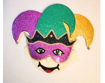 May include: A purple, green, and gold jester mask with a glitter finish. The mask has a black crown, a pink mask with green eyes, and a white face with a smiling mouth.