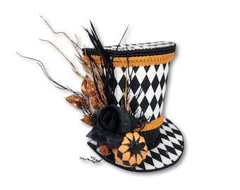 Peut inclure: Un haut-de-forme à motif losanges noir et blanc, orné d'une grande rose en tissu noir, d'une petite citrouille orange, de plumes noires et de brindilles dorées. Le chapeau est bordé d'un cordon tressé noir et doré sur les bords.