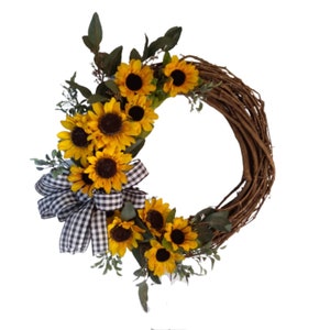 May include: A decorative wreath featuring bright yellow sunflowers with dark brown centers and green leaves. The wreath has a natural brown grapevine base and a black and white checkered bow.