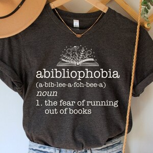 May include: A dark gray t-shirt with a white graphic of an open book with flowers and the text "abibliophobia (a-bib-lee-a-foh-bee-a) noun 1. the fear of running out of books".