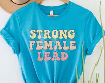 May include: A turquoise blue t-shirt with the text "STRONG FEMALE LEAD" in a retro font style. The text is in yellow, pink, and white.