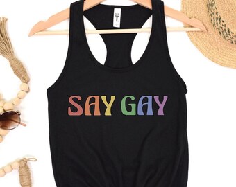May include: Black tank top with the text "SAY GAY" in rainbow colors. The tank top is on a white background with a straw hat and wooden beads.