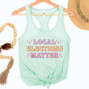 A mint green women’s racerback tank top that says "local elections matter" in purple, yellow, and pink retro text with retro stars