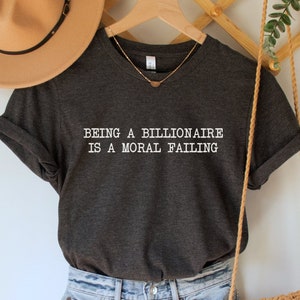 May include: A dark gray t-shirt with the text "Being a billionaire is a moral failing" printed in white. The shirt is hanging on a wooden hanger with a straw hat and a woven bag.
