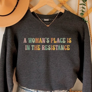 May include: A dark gray sweatshirt with the text "A woman's place is in the resistance" in colorful letters. The sweatshirt is hanging on a wooden hanger with a straw hat and a macrame bag.
