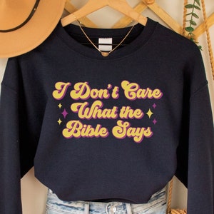 May include: Navy blue sweatshirt with the text "I Don't Care What the Bible Says" in yellow and purple retro lettering with stars. The sweatshirt is hanging on a wooden hanger with a brown woven bag and a hat.