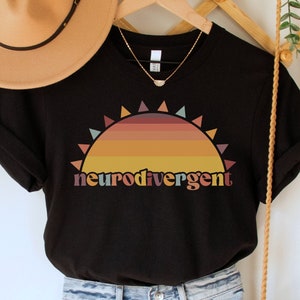 May include: Black t-shirt with a retro sunset graphic and the text "neurodivergent". The shirt is styled with denim shorts and a brown woven bag.