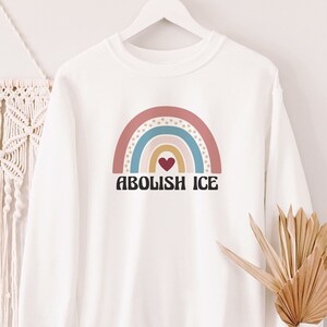 May include: White sweatshirt with a rainbow graphic and the text "Abolish ICE" in black letters. The rainbow is made up of pink, blue, yellow, and orange stripes with a red heart in the center.
