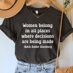 May include: A dark gray t-shirt with the quote "Women belong in all places where decisions are being made -Ruth Bader Ginsburg" printed in white.