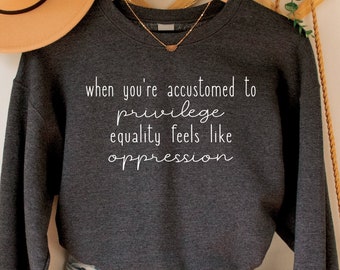 Puede incluir: Sudadera gris oscuro con el texto "when you're accustomed to privilege equality feels like oppression" en blanco. La sudadera está colgada en una percha de madera con un collar dorado. También se ven unos pantalones cortos vaqueros azul claro y un sombrero color canela.