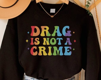 May include: Black sweatshirt with the text "DRAG IS NOT A CRIME" in rainbow colors. The sweatshirt is paired with denim shorts and a woven bag. A tan hat hangs on the wall above the sweatshirt.