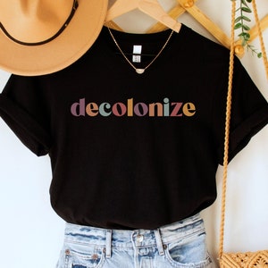 May include: A black t-shirt with the word "decolonize" printed in rainbow colors. The shirt is hanging on a wooden rack with a straw bag and a brown hat.