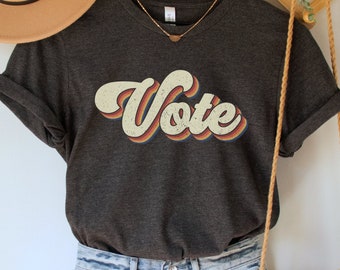 Pode incluir: Uma camiseta cinza escura com um gráfico de "Vote" de estilo retrô em laranja, vermelho e amarelo. A camiseta é combinada com shorts jeans azul claro e uma bolsa de crochê marrom.