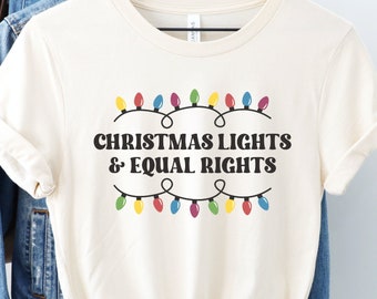 Könnte beinhalten: Cremefarbenes T-Shirt mit dem schwarzen Text "CHRISTMAS LIGHTS & EQUAL RIGHTS". Bunte Lichterketten befinden sich über und unter dem Text. Das T-Shirt hängt an einem Holzkleiderbügel.