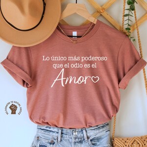 May include: A rust-colored t-shirt with the Spanish text "Lo único más poderoso que el odio es el Amor" is displayed with denim shorts, a tan hat, and a woven bag. The shirt is hanging on a wooden hanger. The outfit is styled on a white background.
