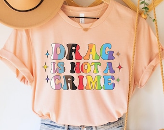 May include: A pink t-shirt with the text "DRAG IS NOT A CRIME" in rainbow letters with stars on either side of the text. The shirt is hanging on a wooden hanger with a brown hat and a brown woven bag.
