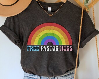 May include: A dark gray t-shirt with a rainbow graphic and the text "FREE PASTOR HUGS". The shirt is paired with light blue denim shorts and a brown crocheted bag.