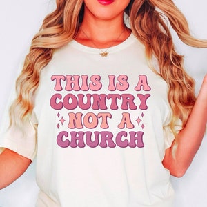 May include: A white t-shirt with the text "This is a country not a church" in pink and peach retro font.