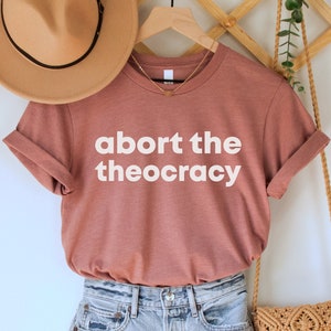 May include: A rust-colored t-shirt with the text "abort the theocracy" in white lettering. The shirt is hanging on a wooden rack with a straw hat and a woven bag.
