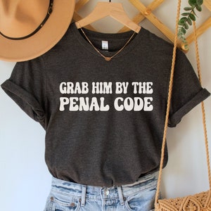 May include: A dark gray t-shirt with the text "Grab Him By The Penal Code" in white retro-style lettering. The shirt is hanging on a wooden hanger and is styled with denim shorts and a brown woven bag.