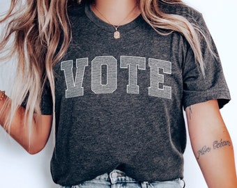 May include: A dark grey t-shirt with the word "VOTE" printed in white on the front. The shirt is being worn by a person with long blonde hair and blue jeans.