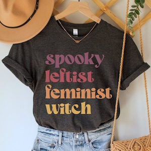 May include: A dark gray t-shirt with the text "spooky leftist feminist witch" in colorful retro font. The shirt is worn with denim shorts and a brown crocheted bag.
