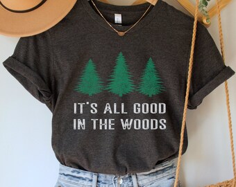 Puede incluir: Una camiseta gris oscuro con un gráfico verde de tres pinos y el texto "It's all good in the woods". La camiseta está colgada en una percha de madera con un sombrero marrón y una bolsa tejida.