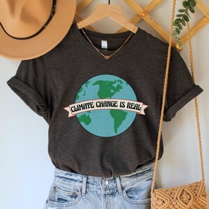 May include: A dark grey t-shirt with a graphic of the Earth and the text "Climate Change is Real" on a banner. The shirt is on a hanger with a brown hat and a woven bag.