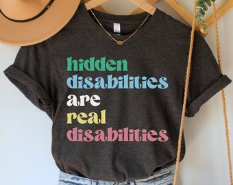 May include: A dark grey t-shirt with the text "hidden disabilities are real disabilities" in colourful letters. The shirt is hanging on a wooden hanger with a straw hat and a crocheted bag.