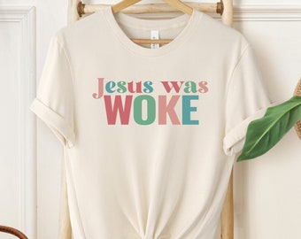 May include: A cream-colored t-shirt with the text "Jesus was WOKE" in colorful letters. The letters are in shades of pink, green, and blue.