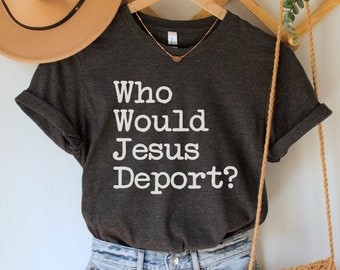 Può includere: Una maglietta grigia scura con la scritta "Who Would Jesus Deport?" stampata in bianco. La maglietta è appesa a un appendiabiti di legno con un cappello marrone e una borsa intrecciata.