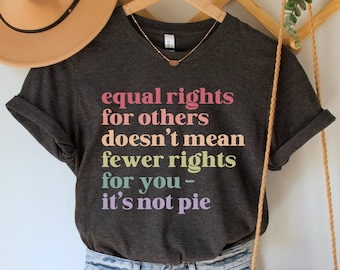 May include: A dark gray t-shirt with the text "equal rights for others doesn't mean fewer rights for you - it's not pie" in rainbow colors. The shirt is worn with light blue denim shorts and a brown crocheted bag.